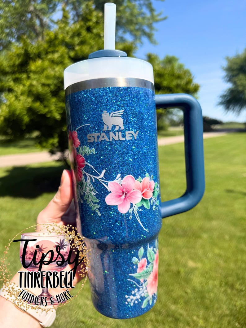 Może przedstawiać: Niebieski kubek Stanley z brokatowym wykończeniem, ozdobiony kwiatowym wzorem z r&oacute;żowymi kwiatami typu hibiskus i zielonymi liśćmi. Kubek posiada ciemnoniebieski uchwyt, białą pokrywkę i przezroczystą słomkę. Logo Stanley jest widoczne w kolorze srebrnym.