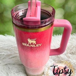 May include: A pink and white ombre Stanley tumbler with a handle and straw. The tumbler has a gold band and the Stanley logo. The text "Tipsy Tinkerbell Tumblers & More" is in a circular design.