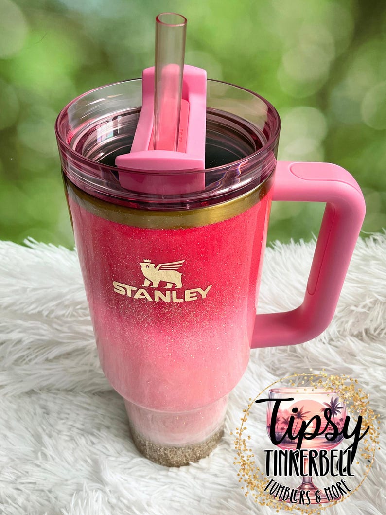 May include: A pink and white ombre Stanley tumbler with a handle and straw. The tumbler has a gold band and the Stanley logo. The text "Tipsy Tinkerbell Tumblers & More" is in a circular design.