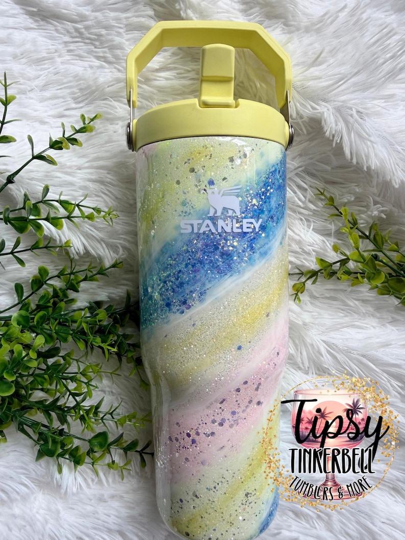 May include: A Stanley tumbler with a pastel swirl design in shades of blue, yellow, pink, and white, accented with glitter. The tumbler has a yellow handle and lid. The Stanley logo is visible on the front.