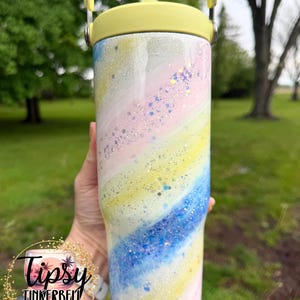 May include: A pastel-colored tumbler with a yellow handle and lid. The tumbler features a swirling design of pink, yellow, and blue with glitter accents. The text "Tipsy Tinkerbell Tumblers & More" is visible.