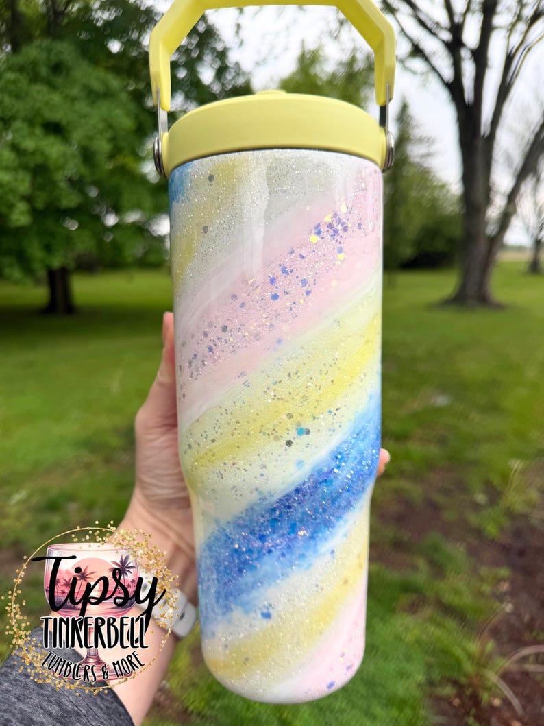May include: A pastel-colored tumbler with a yellow handle and lid. The tumbler features a swirling design of pink, yellow, and blue with glitter accents. The text "Tipsy Tinkerbell Tumblers & More" is visible.