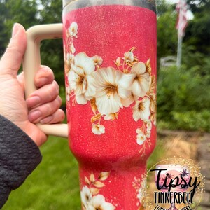 K&ouml;nnte beinhalten: Ein hoher, glitzernder korallenroter Becher mit einem Blumenmuster aus wei&szlig;en und cremefarbenen Hibiskusbl&uuml;ten. Das Trinkgef&auml;&szlig; verf&uuml;gt &uuml;ber einen beigen Henkel, einen durchsichtigen Deckel mit beigem Verschluss und ein rundes Logo mit der Aufschrift Tipsy Tinkerbell Tumblers & More.