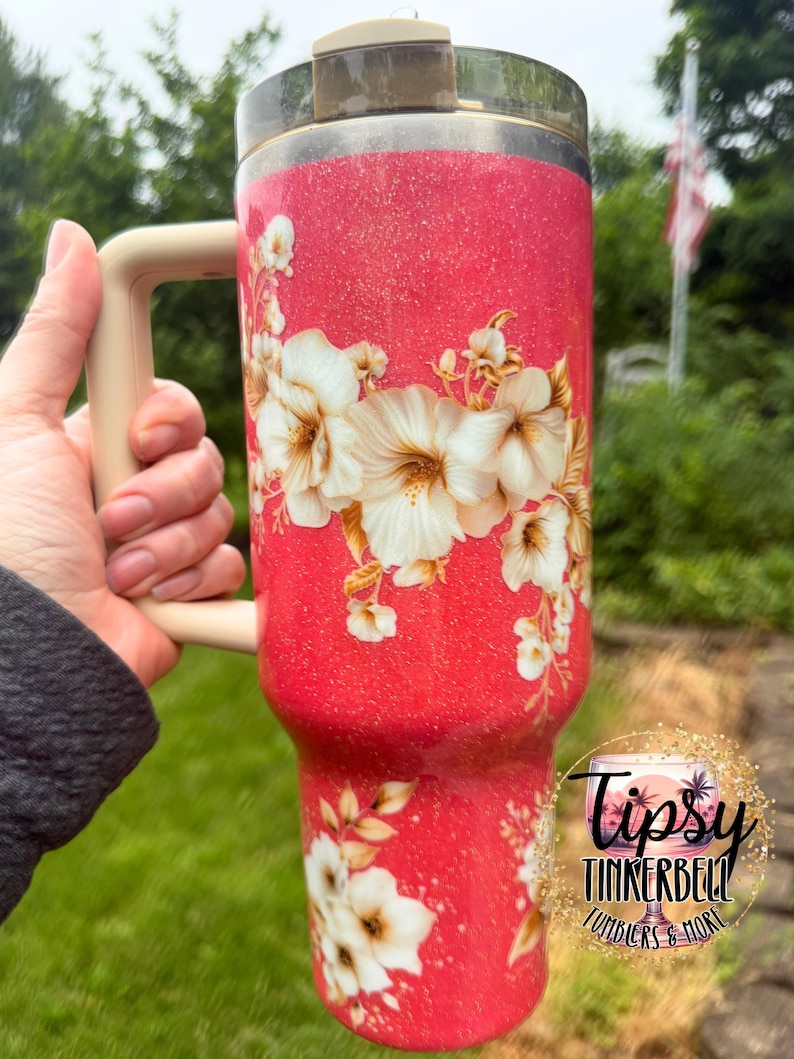 K&ouml;nnte beinhalten: Ein hoher, glitzernder korallenroter Becher mit einem Blumenmuster aus wei&szlig;en und cremefarbenen Hibiskusbl&uuml;ten. Das Trinkgef&auml;&szlig; verf&uuml;gt &uuml;ber einen beigen Henkel, einen durchsichtigen Deckel mit beigem Verschluss und ein rundes Logo mit der Aufschrift Tipsy Tinkerbell Tumblers & More.