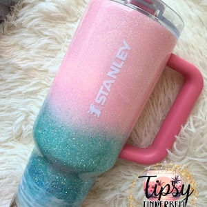 May include: A Stanley tumbler featuring a glittery ombre finish that transitions from light pink at the top to teal and white waves in the middle, ending with a gold glitter base. The cup has a pink handle, a clear lid, and a pink straw.