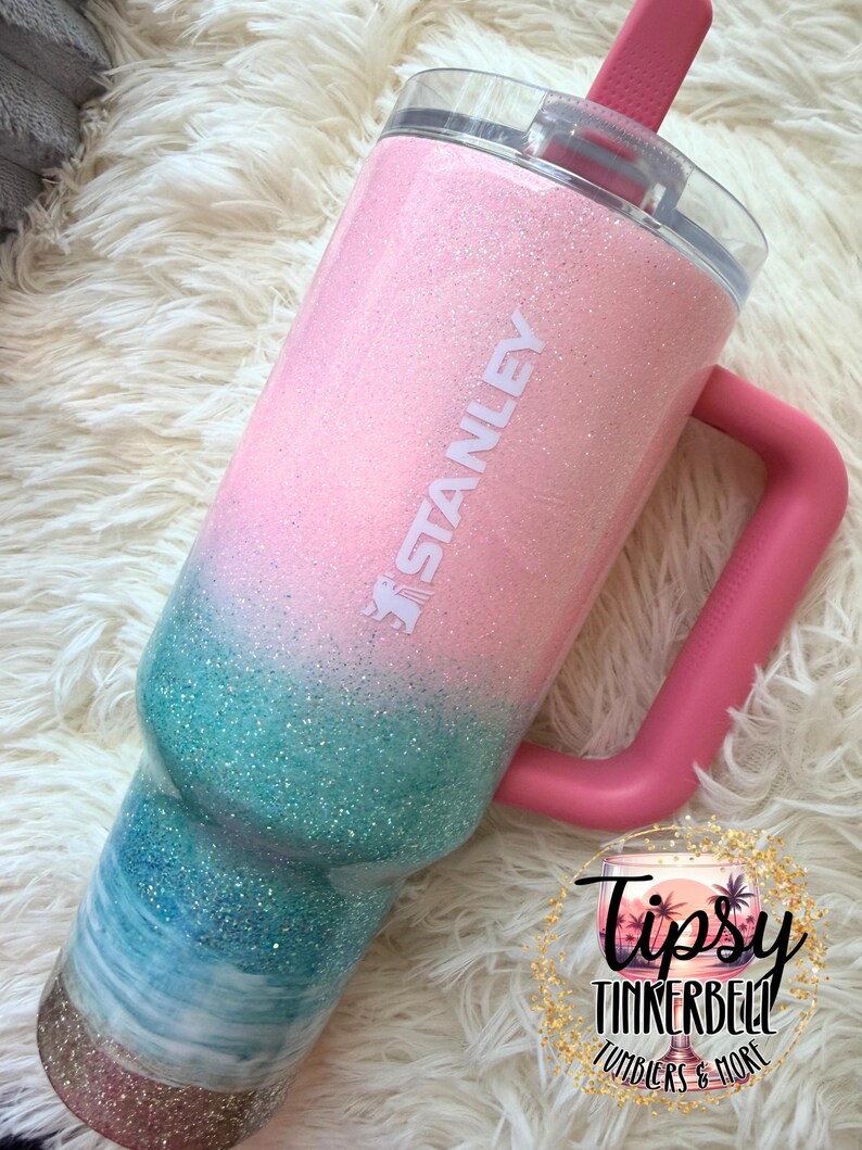 May include: A Stanley tumbler featuring a glittery ombre finish that transitions from light pink at the top to teal and white waves in the middle, ending with a gold glitter base. The cup has a pink handle, a clear lid, and a pink straw.