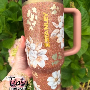 May include: A copper-coloured glitter tumbler with a handle and a dark lid. The tumbler features white floral illustrations, gold-coloured leopard print accents, and the word STANLEY printed vertically in gold lettering on the side.