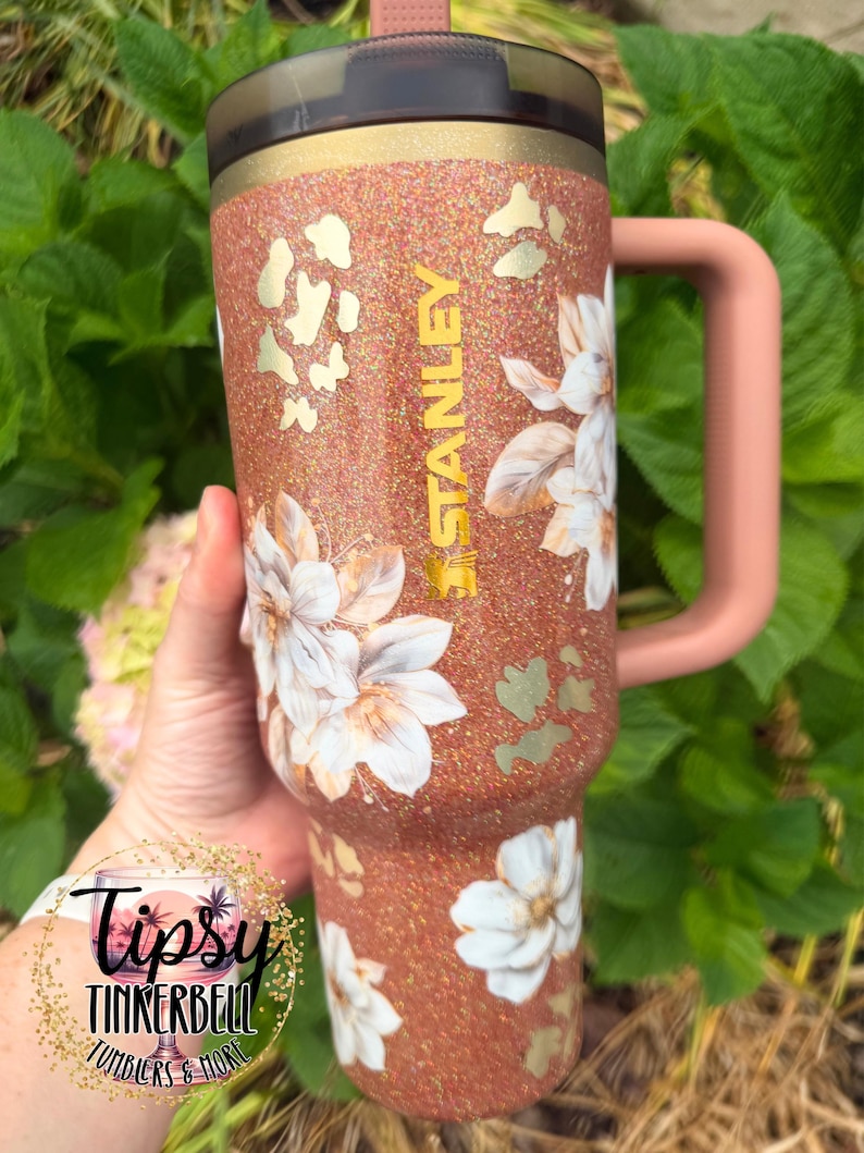 May include: A copper-coloured glitter tumbler with a handle and a dark lid. The tumbler features white floral illustrations, gold-coloured leopard print accents, and the word STANLEY printed vertically in gold lettering on the side.