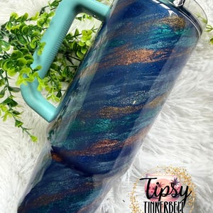 Puede incluir: Un vaso grande y aislado con un asa y tapa de color turquesa. El vaso presenta un dise&ntilde;o arremolinado de purpurina azul oscuro, turquesa y bronce. El vaso est&aacute; decorado con el logotipo "Tipsy Tinkerbell Tumblers & More".