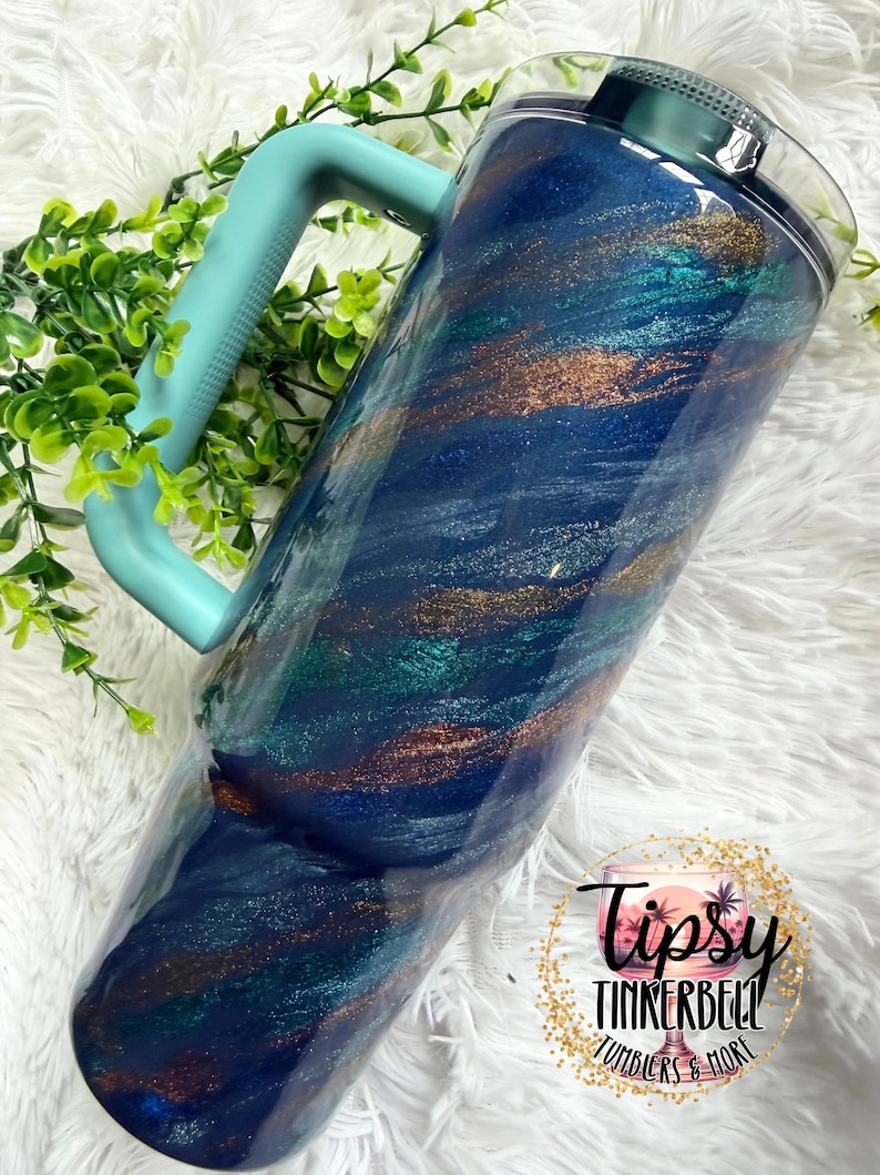 Puede incluir: Un vaso grande y aislado con un asa y tapa de color turquesa. El vaso presenta un dise&ntilde;o arremolinado de purpurina azul oscuro, turquesa y bronce. El vaso est&aacute; decorado con el logotipo "Tipsy Tinkerbell Tumblers & More".