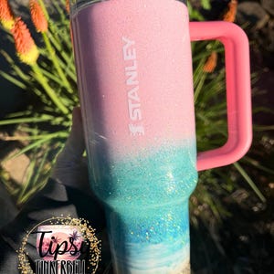 May include: A Stanley tumbler featuring a pink top section that transitions into a glittery teal and blue ombre design, ending in a sandy-colored base. The cup includes a pink handle, a clear lid with a pink straw, and the word STANLEY embossed vertically.