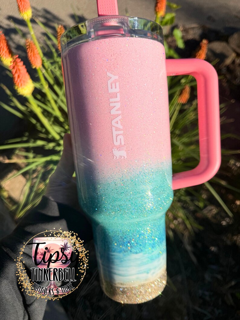 May include: A Stanley tumbler featuring a pink top section that transitions into a glittery teal and blue ombre design, ending in a sandy-colored base. The cup includes a pink handle, a clear lid with a pink straw, and the word STANLEY embossed vertically.