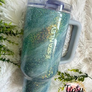 May include: A teal and blue glitter Stanley tumbler with a handle and a clear lid. The tumbler has a marbled effect with gold glitter accents and the Stanley logo. The tumbler is on a white fluffy surface.