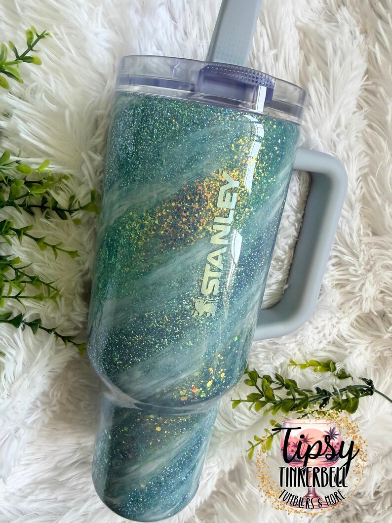 May include: A teal and blue glitter Stanley tumbler with a handle and a clear lid. The tumbler has a marbled effect with gold glitter accents and the Stanley logo. The tumbler is on a white fluffy surface.
