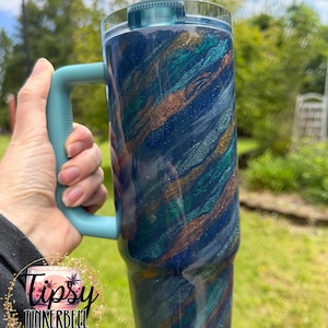 Puede incluir: Un vaso grande y aislado con un asa y tapa color turquesa. El cuerpo del vaso es azul oscuro con vetas de turquesa, dorado y purpurina plateada. El vaso se sostiene frente a un fondo verde.