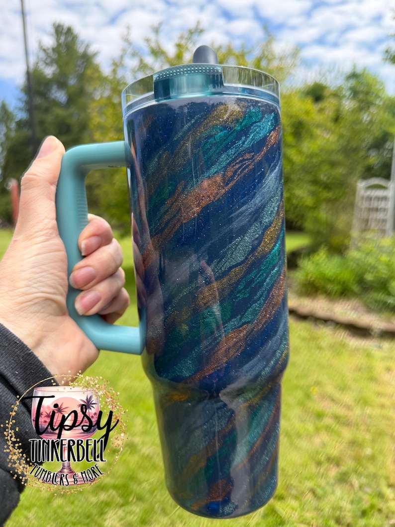 Puede incluir: Un vaso grande y aislado con un asa y tapa color turquesa. El cuerpo del vaso es azul oscuro con vetas de turquesa, dorado y purpurina plateada. El vaso se sostiene frente a un fondo verde.