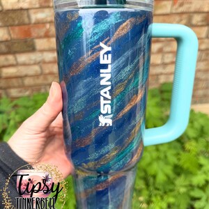 Puede incluir: Un vaso Stanley azul con asa y tapa turquesa. El vaso presenta un dise&ntilde;o brillante y arremolinado en tonos azules, turquesas y dorados. La palabra "STANLEY" est&aacute; impresa en blanco. El vaso es sostenido por una persona.