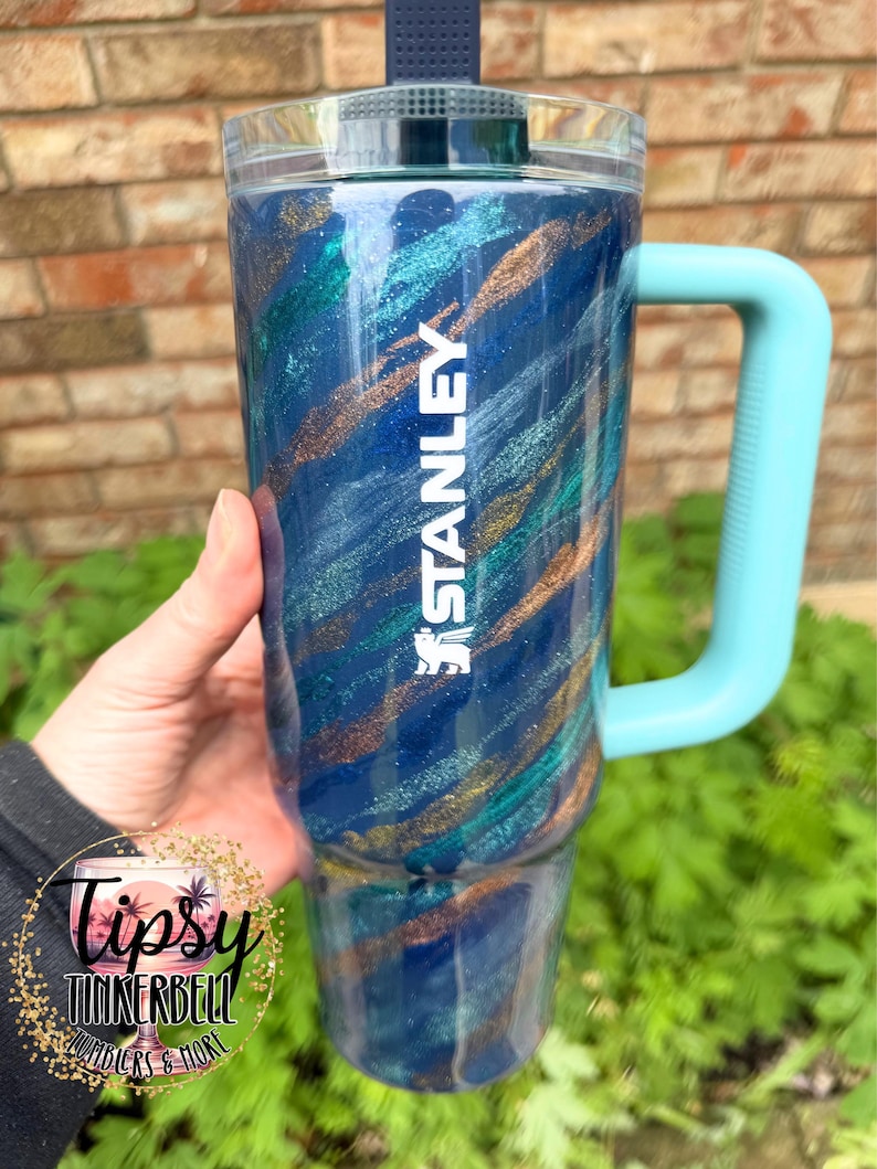 Puede incluir: Un vaso Stanley azul con asa y tapa turquesa. El vaso presenta un dise&ntilde;o brillante y arremolinado en tonos azules, turquesas y dorados. La palabra "STANLEY" est&aacute; impresa en blanco. El vaso es sostenido por una persona.