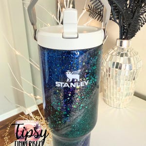 May include: A Stanley tumbler featuring a white lid and a gray handle, with a body covered in a dark blue, green, and silver glitter swirl pattern. The white Stanley logo with a bear icon is printed on the front. A circular logo reads Tipsy Tinkerbell.