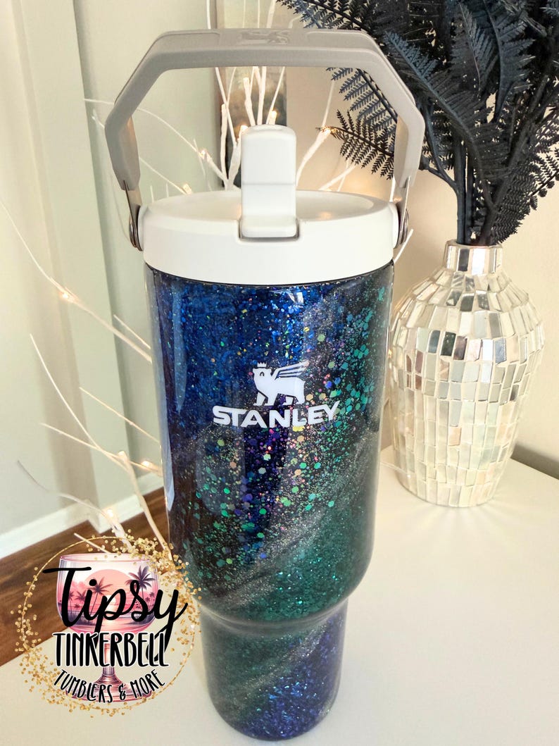 May include: A Stanley tumbler featuring a white lid and a gray handle, with a body covered in a dark blue, green, and silver glitter swirl pattern. The white Stanley logo with a bear icon is printed on the front. A circular logo reads Tipsy Tinkerbell.