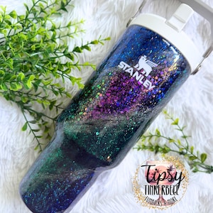 May include: A Stanley brand tumbler featuring a glossy finish with a galaxy-inspired design in shades of deep blue, purple, and green, accented with iridescent glitter. The tumbler has a white lid with a handle and the white Stanley logo printed.