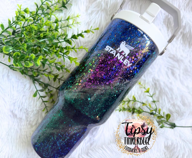May include: A Stanley brand tumbler featuring a glossy finish with a galaxy-inspired design in shades of deep blue, purple, and green, accented with iridescent glitter. The tumbler has a white lid with a handle and the white Stanley logo printed.