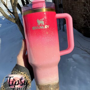May include: A pink Stanley tumbler with a handle and straw. The tumbler has a glitter ombre effect, transitioning from red to pink to white, with a gold glitter base. The Stanley logo is visible on the front. The tumbler is a product of Tipsy Tinkerbell Tumblers & More.
