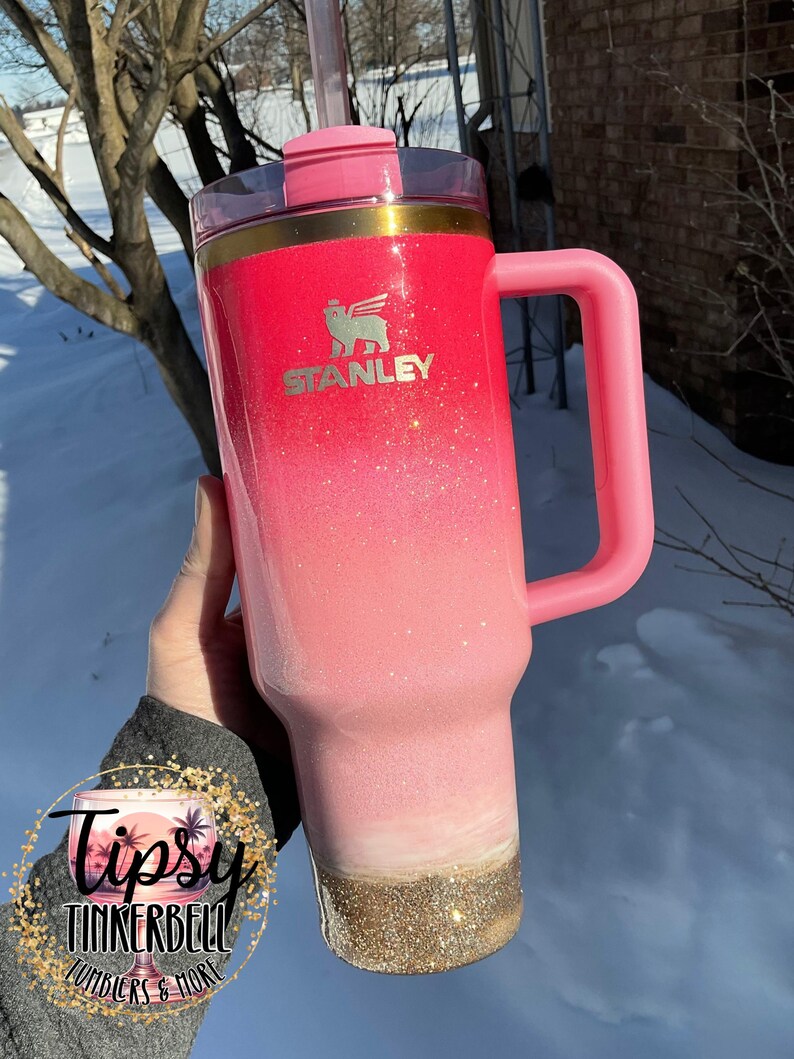 May include: A pink Stanley tumbler with a handle and straw. The tumbler has a glitter ombre effect, transitioning from red to pink to white, with a gold glitter base. The Stanley logo is visible on the front. The tumbler is a product of Tipsy Tinkerbell Tumblers & More.