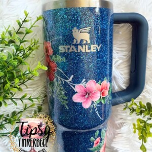 Może przedstawiać: Niebieski kubek Stanley z brokatowym wykończeniem i kwiatowym wzorem z r&oacute;żowymi i czerwonymi kwiatami hibiskusa oraz zielonymi liśćmi. Kubek posiada ciemnoniebieski uchwyt, przezroczystą pokrywkę z ciemnoniebieską zatyczką oraz logo Stanley.