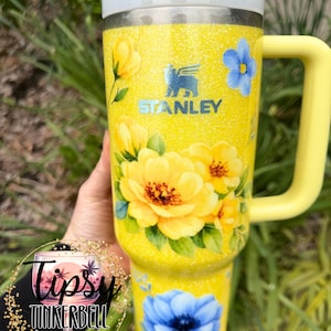 May include: A yellow Stanley tumbler with a floral design featuring yellow and blue flowers. The tumbler has a yellow handle and a white lid with a straw. The tumbler is decorated with glitter and the Stanley logo.