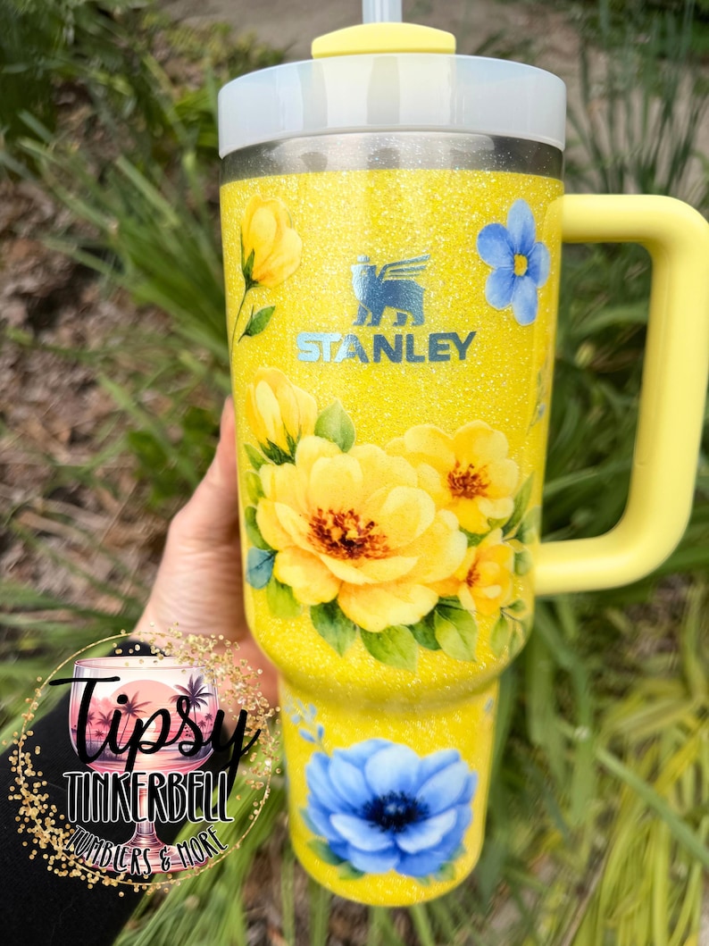 May include: A yellow Stanley tumbler with a floral design featuring yellow and blue flowers. The tumbler has a yellow handle and a white lid with a straw. The tumbler is decorated with glitter and the Stanley logo.