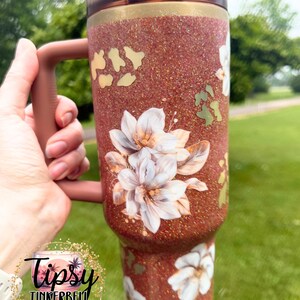 May include: A copper-coloured glitter tumbler with a handle and a dark translucent lid. The surface features a pattern of white floral illustrations and gold-toned leopard print spots. A circular logo with the text Tipsy Tinkerbell Tumblers & More.