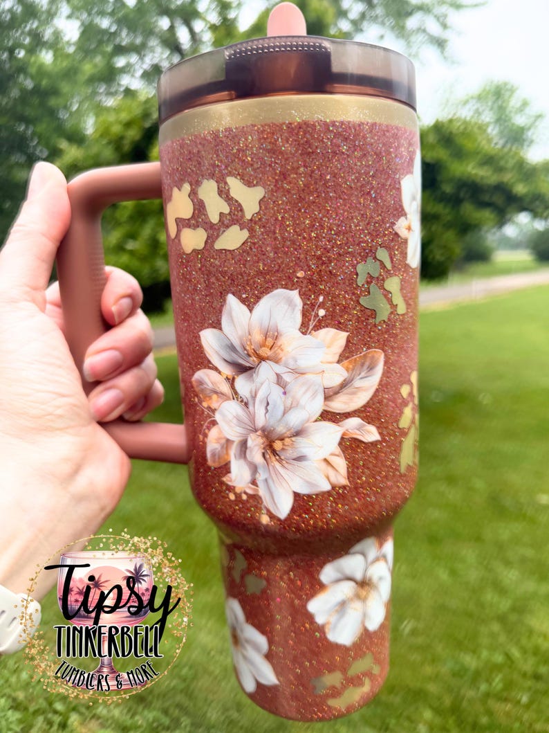 May include: A copper-coloured glitter tumbler with a handle and a dark translucent lid. The surface features a pattern of white floral illustrations and gold-toned leopard print spots. A circular logo with the text Tipsy Tinkerbell Tumblers & More.
