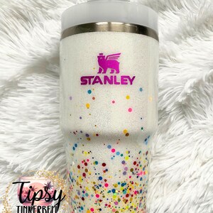 Puede incluir: Un vaso blanco con purpurina y un dise&ntilde;o de confeti colorido. El vaso presenta el logotipo "Stanley" en rosa y una tapa blanca con pajita. La mitad inferior est&aacute; cubierta de confeti colorido.