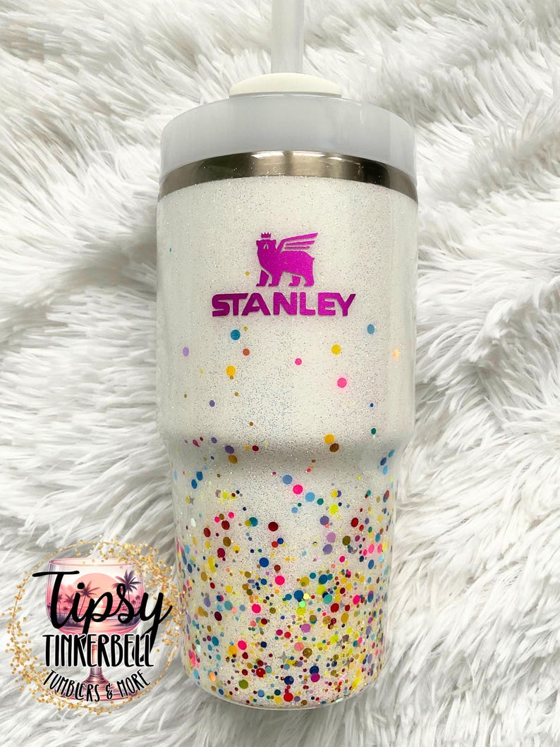 Puede incluir: Un vaso blanco con purpurina y un dise&ntilde;o de confeti colorido. El vaso presenta el logotipo "Stanley" en rosa y una tapa blanca con pajita. La mitad inferior est&aacute; cubierta de confeti colorido.
