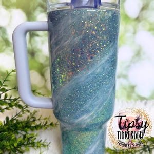 May include: A glittery blue tumbler with a white handle and lid. The tumbler has a clear plastic lid and a white handle. The body of the tumbler is covered in blue glitter with white swirls. The tumbler has the text "Tipsy Tinkerbell Tumblers & More".