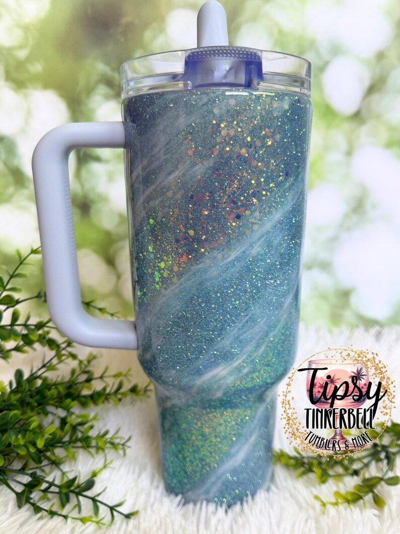 May include: A glittery blue tumbler with a white handle and lid. The tumbler has a clear plastic lid and a white handle. The body of the tumbler is covered in blue glitter with white swirls. The tumbler has the text "Tipsy Tinkerbell Tumblers & More".