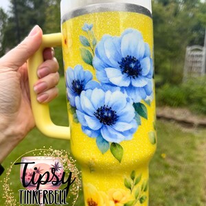 May include: A yellow glitter tumbler with a handle, featuring blue and yellow floral designs. The tumbler has a white lid and a yellow handle. The text "Tipsy Tinkerbell Tumblers & More" is visible on the tumbler.