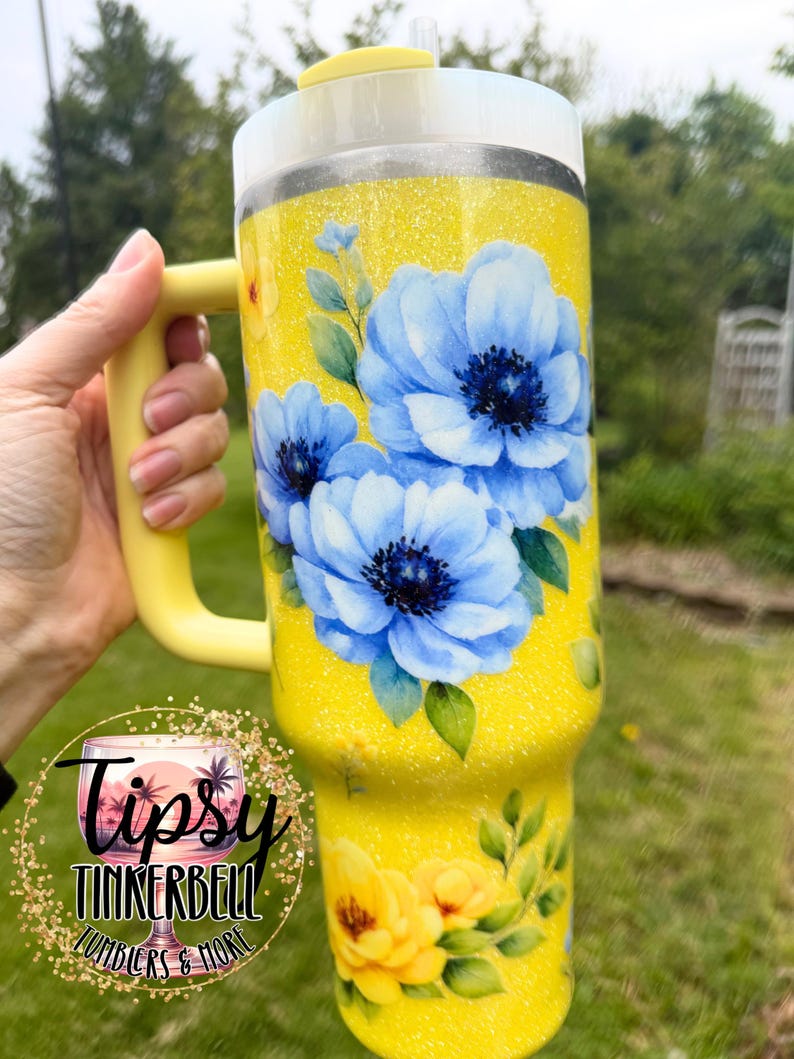 May include: A yellow glitter tumbler with a handle, featuring blue and yellow floral designs. The tumbler has a white lid and a yellow handle. The text "Tipsy Tinkerbell Tumblers & More" is visible on the tumbler.