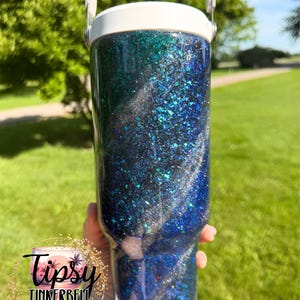 May include: A cylindrical tumbler with a white lid and handle, featuring a swirling glitter finish in shades of deep blue, teal, and black. The surface is covered in reflective, multi-sized glitter particles that create a textured, shimmering appearance.