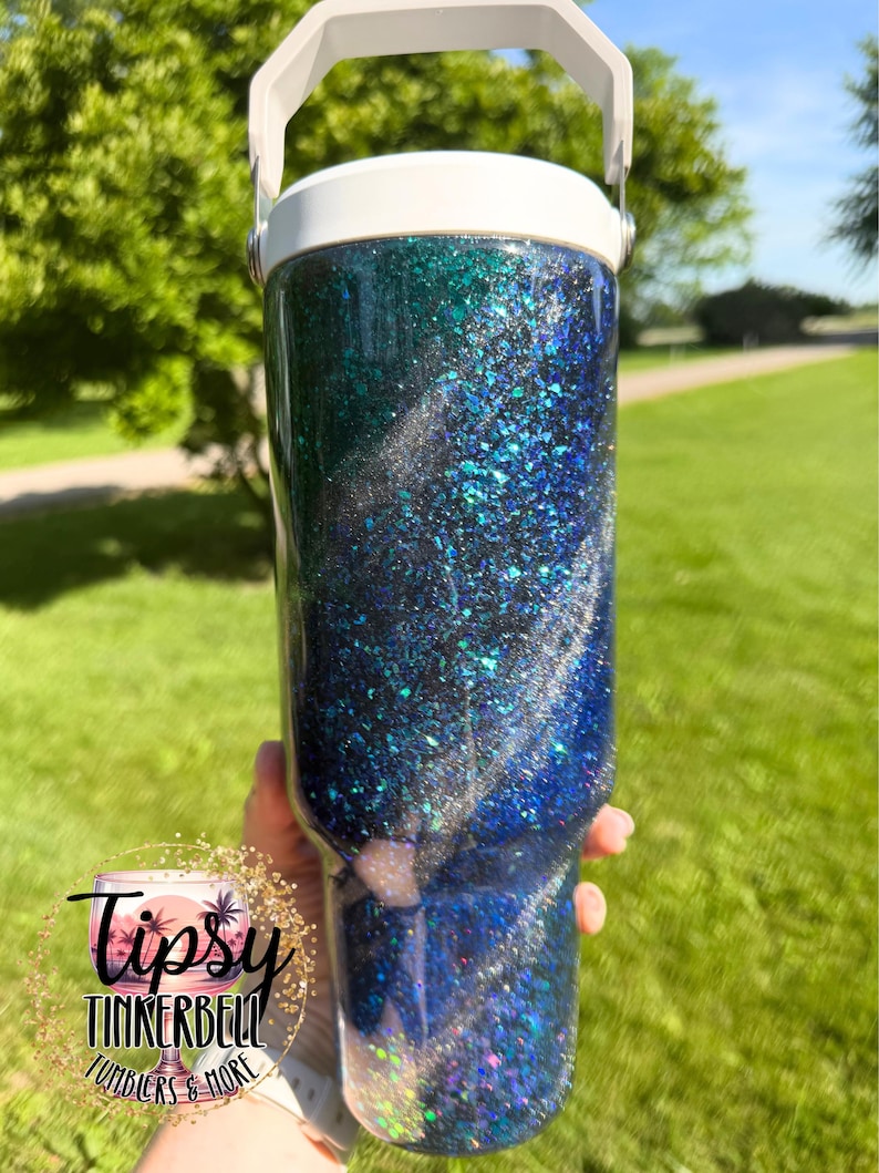 May include: A cylindrical tumbler with a white lid and handle, featuring a swirling glitter finish in shades of deep blue, teal, and black. The surface is covered in reflective, multi-sized glitter particles that create a textured, shimmering appearance.