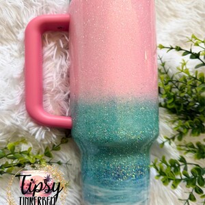 May include: A stainless steel tumbler with a handle and a clear lid, featuring a glittery ombre design that transitions from light pink at the top to teal, blue, and white ocean waves, ending with a gold glitter base. Includes a circular logo.