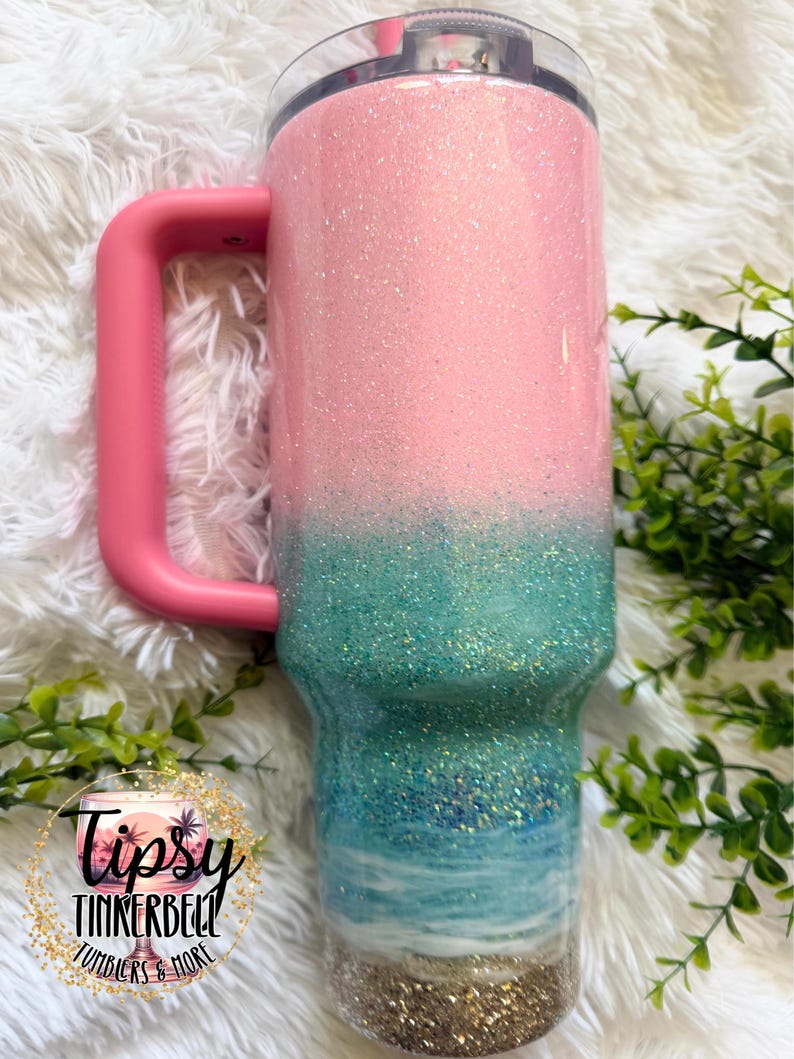 May include: A stainless steel tumbler with a handle and a clear lid, featuring a glittery ombre design that transitions from light pink at the top to teal, blue, and white ocean waves, ending with a gold glitter base. Includes a circular logo.
