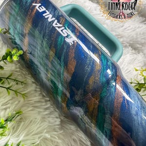 Puede incluir: Un vaso Stanley grande y aislado con un asa y tapa color turquesa. El vaso presenta un dise&ntilde;o arremolinado de pintura met&aacute;lica azul, verde y dorada. La palabra "STANLEY" est&aacute; impresa en blanco. La imagen tambi&eacute;n incluye el texto "Tipsy Tinkerbell Tumblers & More".
