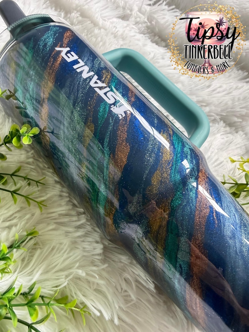 Puede incluir: Un vaso Stanley grande y aislado con un asa y tapa color turquesa. El vaso presenta un dise&ntilde;o arremolinado de pintura met&aacute;lica azul, verde y dorada. La palabra "STANLEY" est&aacute; impresa en blanco. La imagen tambi&eacute;n incluye el texto "Tipsy Tinkerbell Tumblers & More".