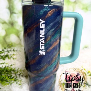 Puede incluir: Un vaso grande y aislado con un dise&ntilde;o en espiral azul, verde azulado y bronce. El vaso tiene un asa azul claro y la palabra "STANLEY" en blanco. El vaso tiene una tapa negra y una pajita. El vaso es de 1,18 litros.