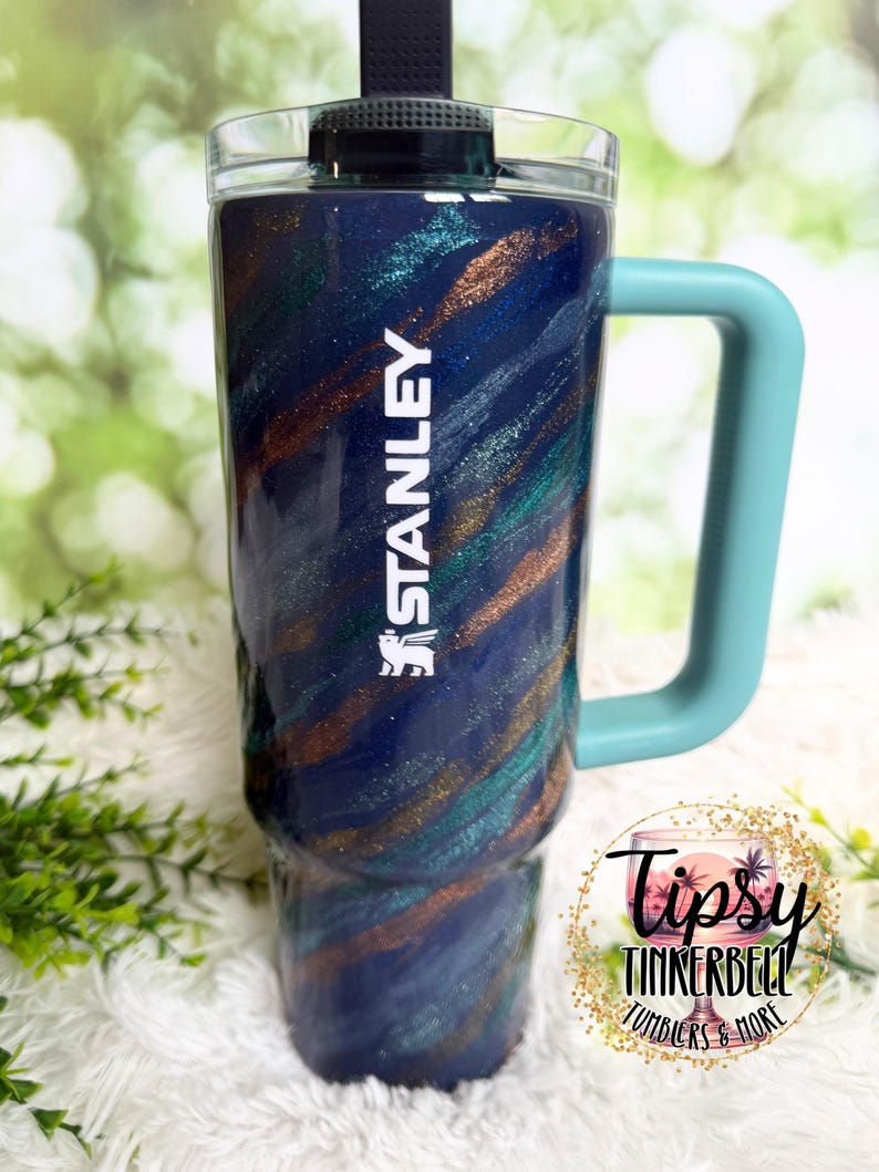 Puede incluir: Un vaso grande y aislado con un dise&ntilde;o en espiral azul, verde azulado y bronce. El vaso tiene un asa azul claro y la palabra "STANLEY" en blanco. El vaso tiene una tapa negra y una pajita. El vaso es de 1,18 litros.