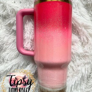 May include: A pink ombre tumbler with a handle, featuring a glittery finish. The tumbler has a clear lid and a gold band. The bottom section is a gold glitter. The text "Tipsy Tinkerbell Tumblers & More" is visible.