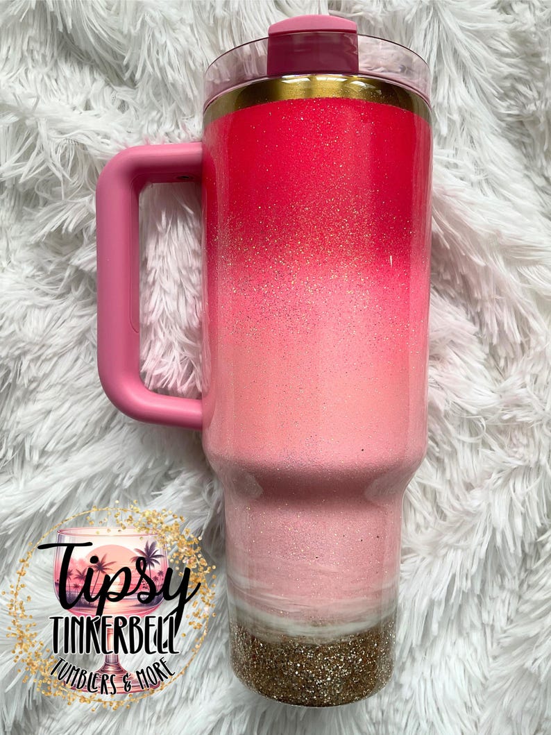 May include: A pink ombre tumbler with a handle, featuring a glittery finish. The tumbler has a clear lid and a gold band. The bottom section is a gold glitter. The text "Tipsy Tinkerbell Tumblers & More" is visible.