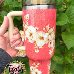 K&ouml;nnte beinhalten: Ein korallenfarbener Becher mit Glitzer-Finish, verziert mit einem wei&szlig;en Blumenmuster, einem beigen Henkel und einem dunkel durchscheinenden Deckel mit Strohhalm. Der Becher zeigt ein Logo mit dem Text Tipsy Tinkerbell Tumblers & More.